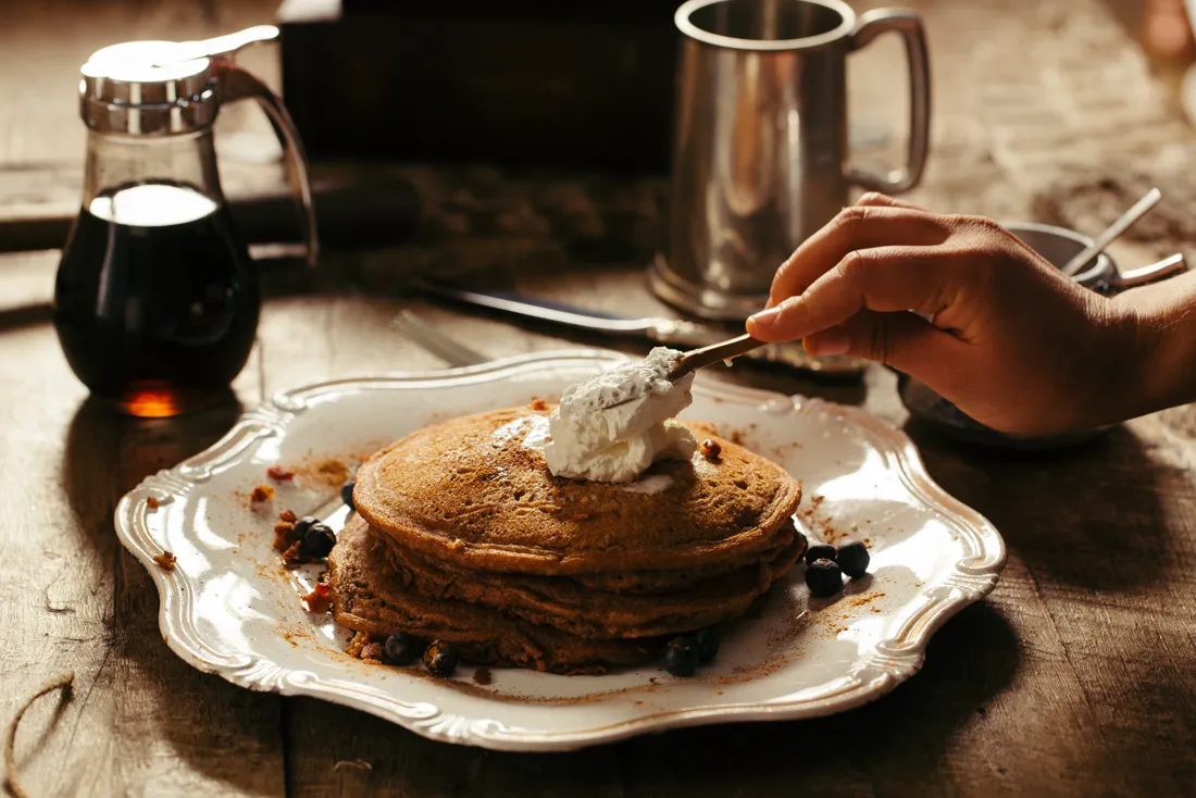 Stack of pancakes with cream and blueberries on a breakfast table, syrup jug and mug behind